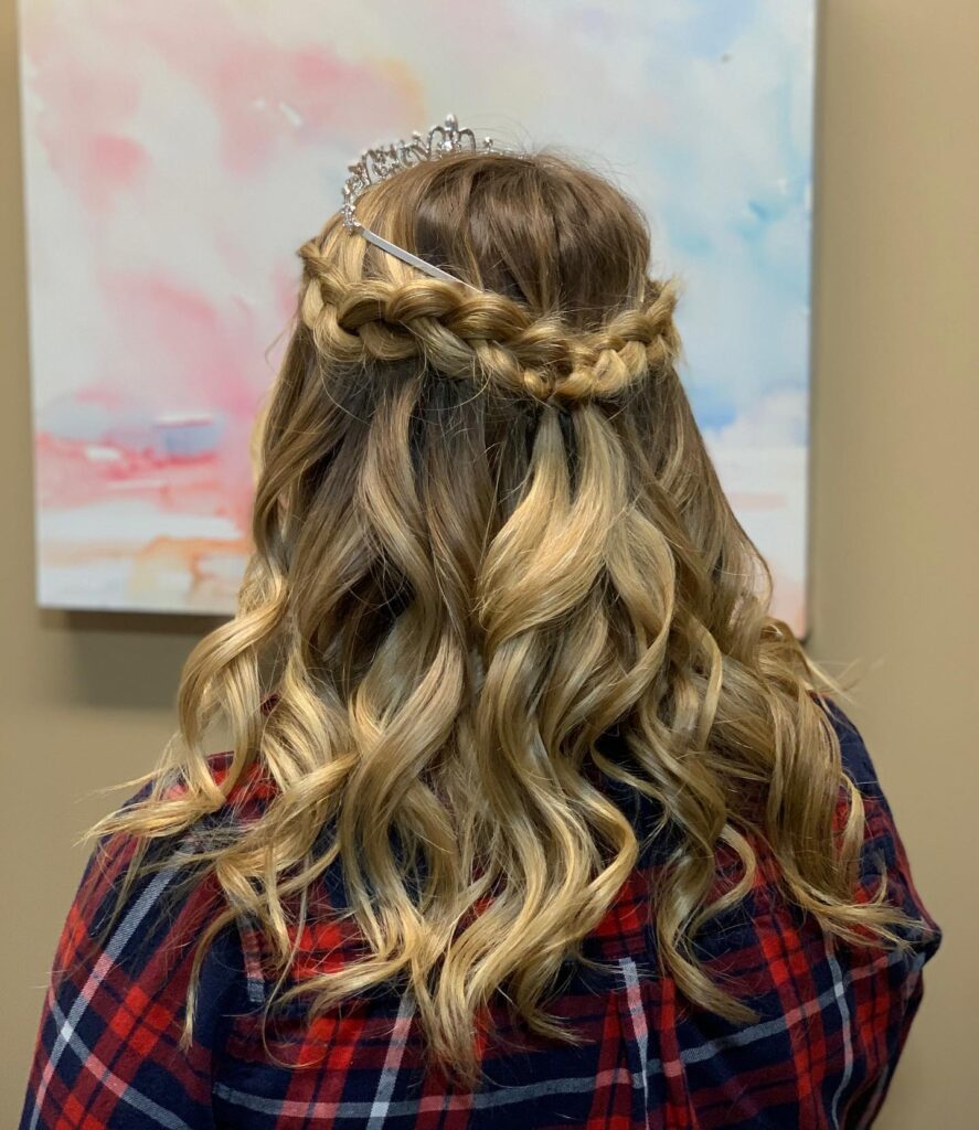 A woman with medium-length curly blonde hair is styled with a thick horizontal braid across the back and a silver tiara on top.