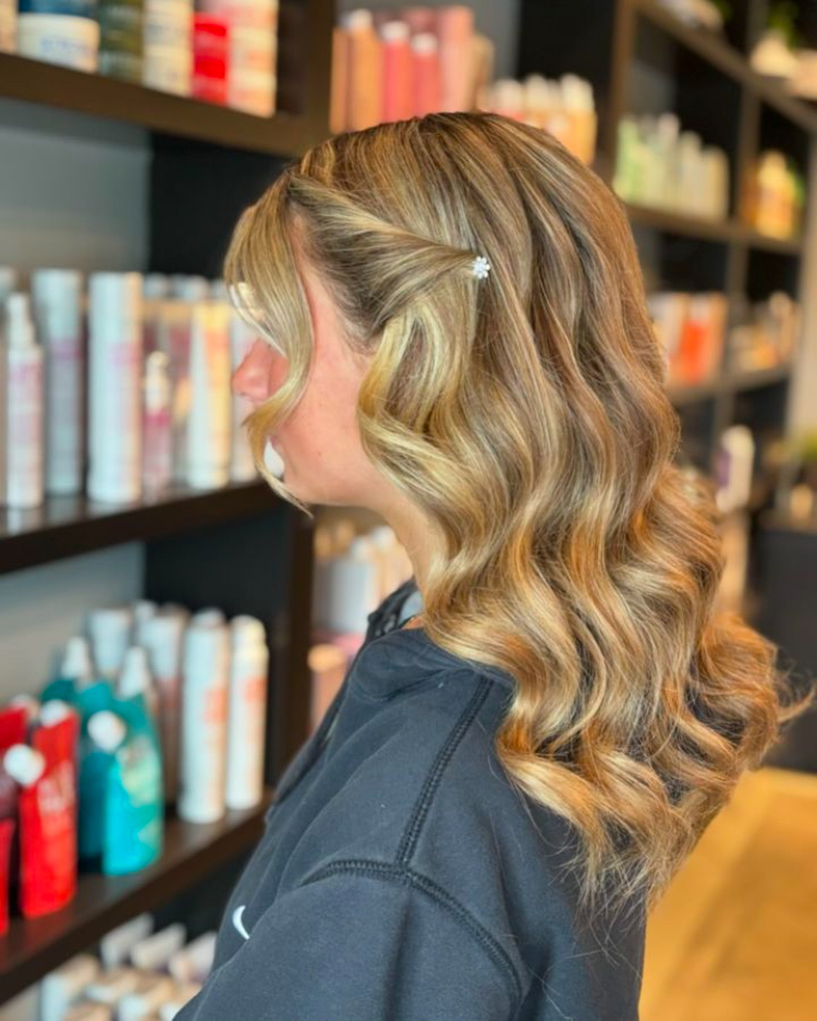 A woman with medium-length wavy caramel-blonde hair has one side pinned back with a small white flower clip.