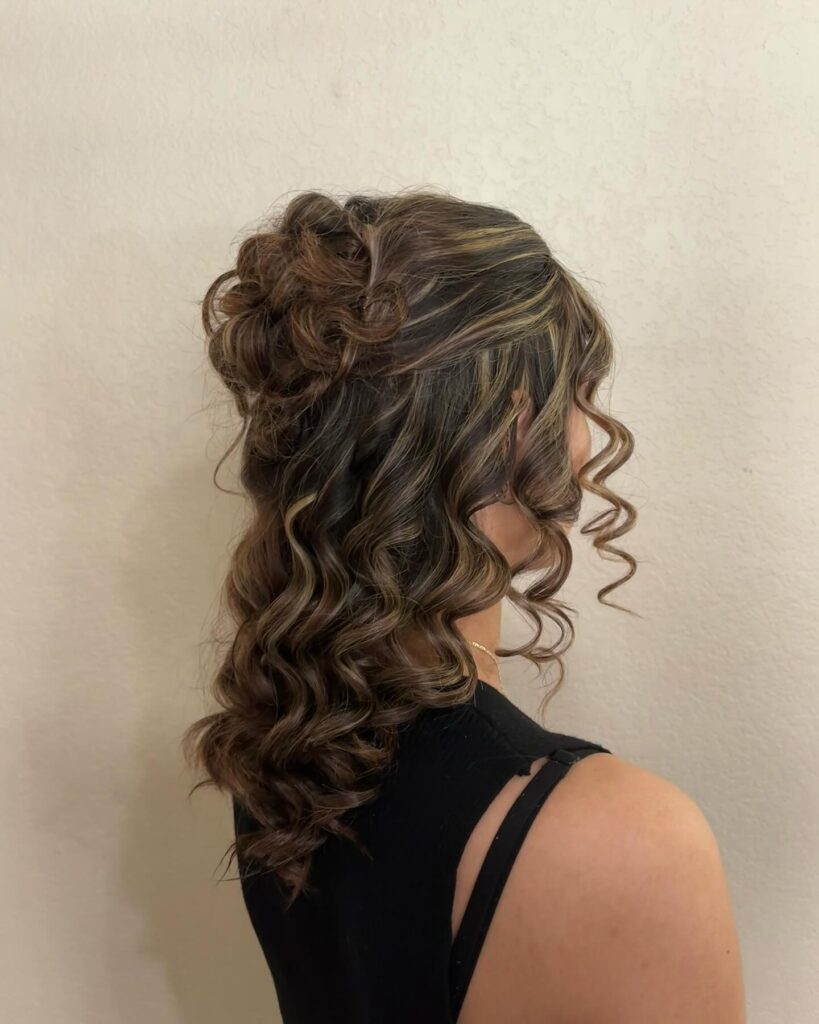 A side profile of a woman with medium-length dark brown hair and blonde highlights styled in a messy half-up bun with tight ringlet curls.