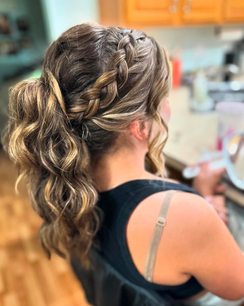 A woman with wavy brunette hair and blonde highlights styled in a side Dutch braid that leads into a curly ponytail.