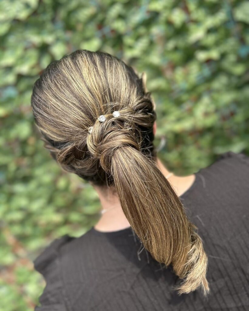 A woman with highlighted brown hair styled in a sleek low ponytail with a hair wrap around the base and three small crystal hairpins.