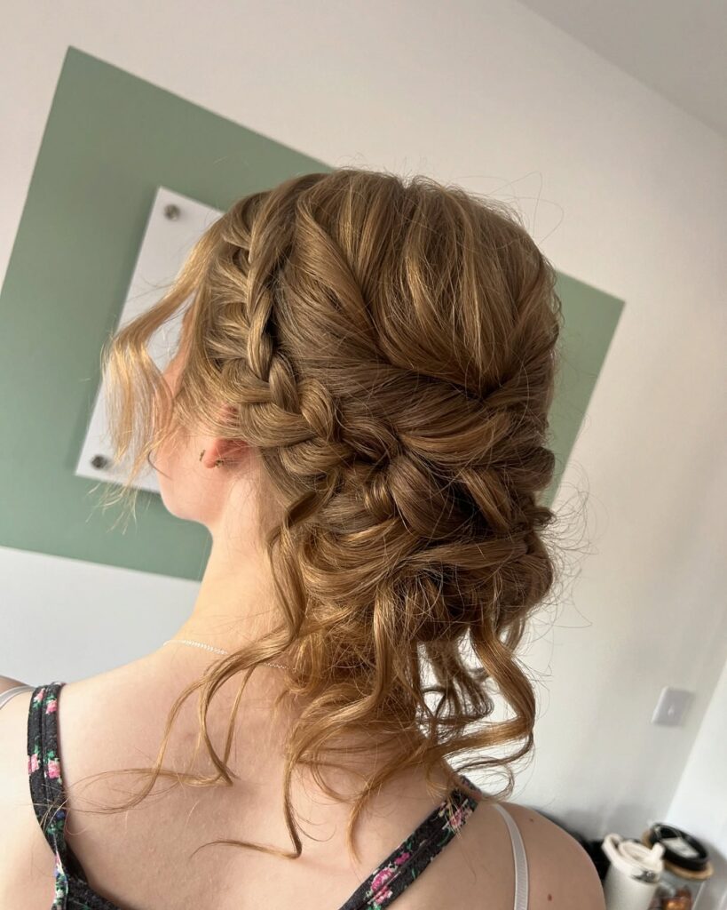 A woman with medium brown hair styled in a thick side braid that leads into a messy low updo with loose curly tendrils.
