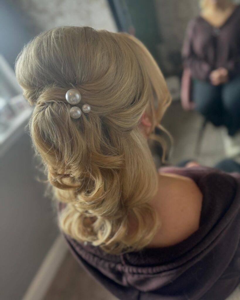 A woman with medium-length blonde hair styled in a voluminous bouffant half-updo accented with three white pearls and soft curls.