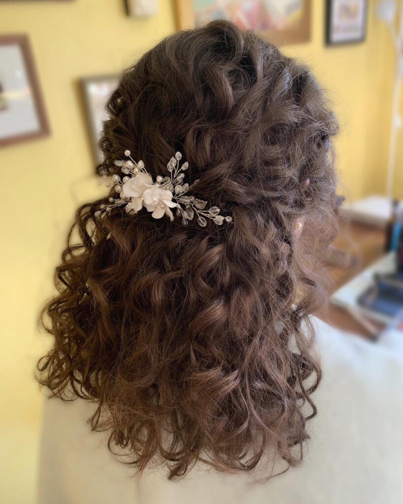 A woman with shoulder-length voluminous curly brown hair is styled in a half-up look, accented by a white floral and crystal hair vine.