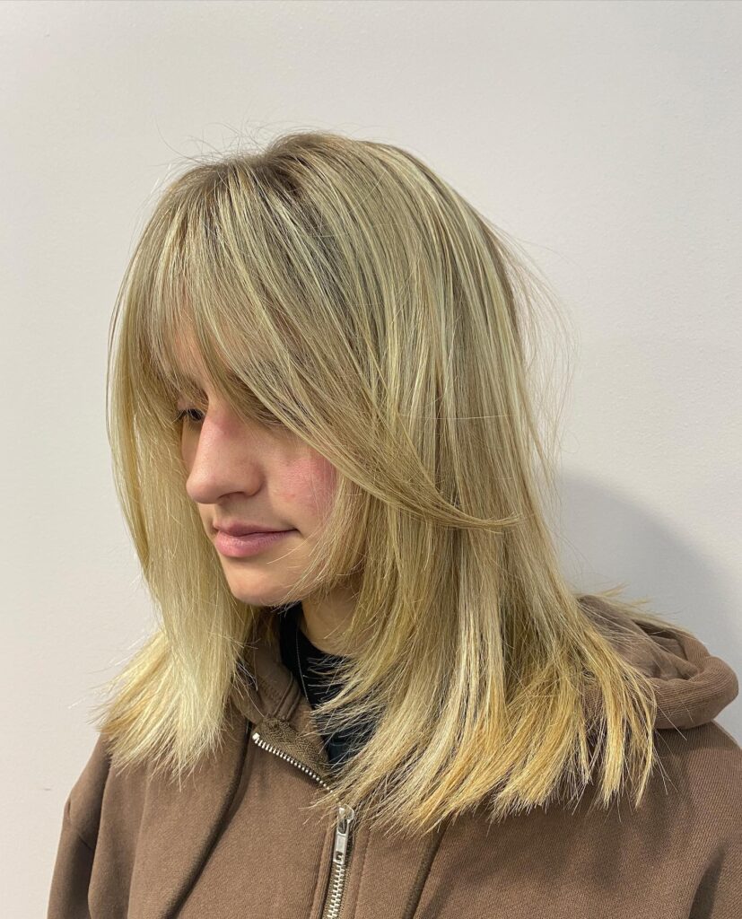 A person with medium-length straight sandy blonde hair featuring choppy face-framing layers and long, wispy bangs.