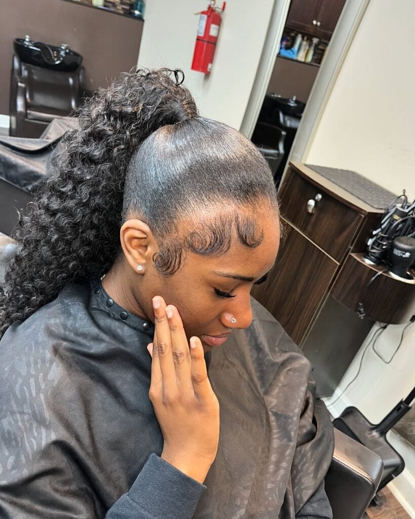 A young woman with sleek dark hair pulled into a high ponytail featuring dense, voluminous curls and styled baby hairs.