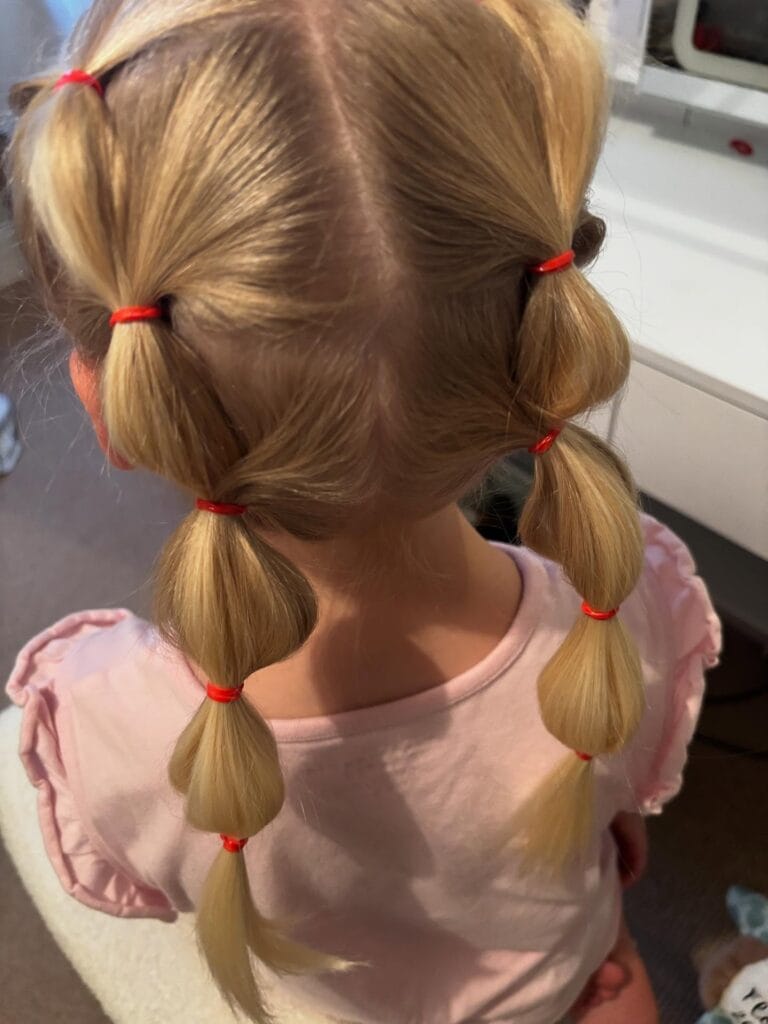 A young girl with blonde hair styled into two bubble pigtails held by bright red elastic bands.