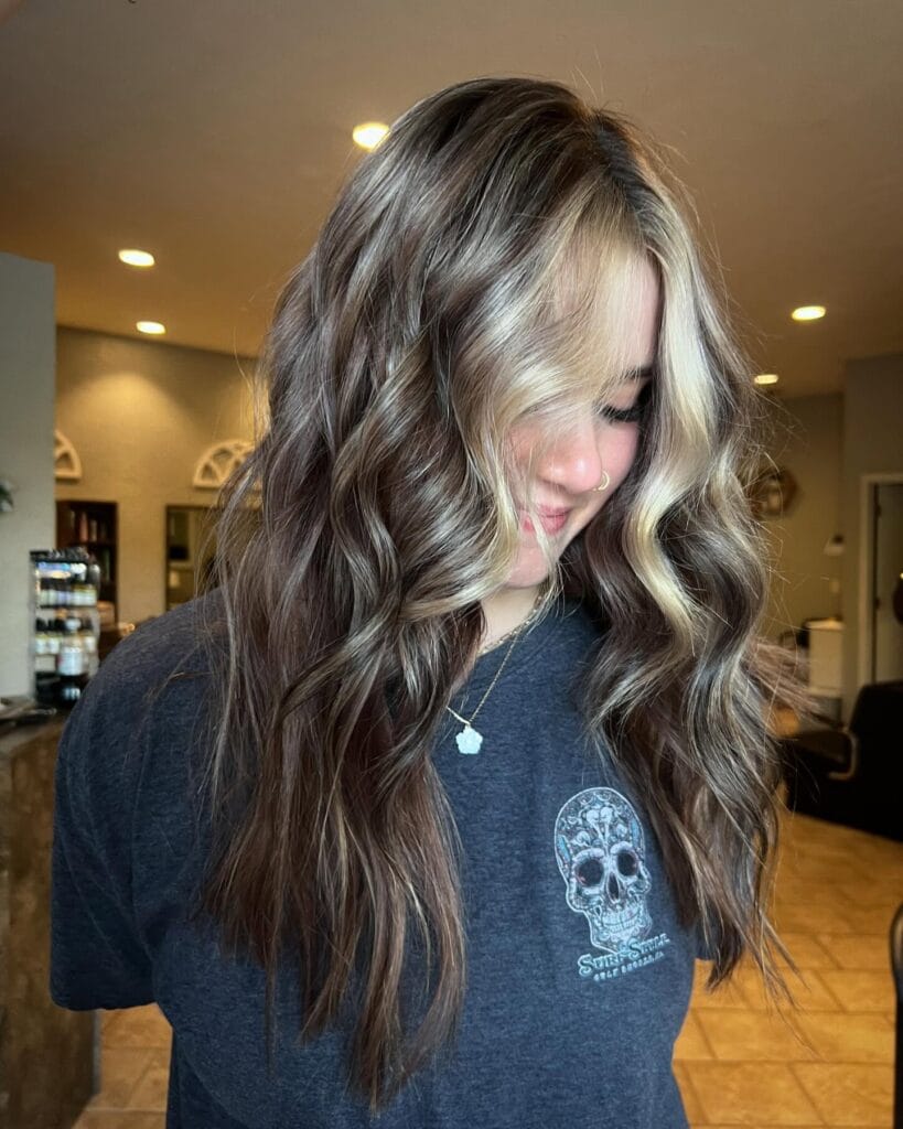 A young woman with long, wavy, brunette hair featuring bright blonde money piece highlights and beachy texture.