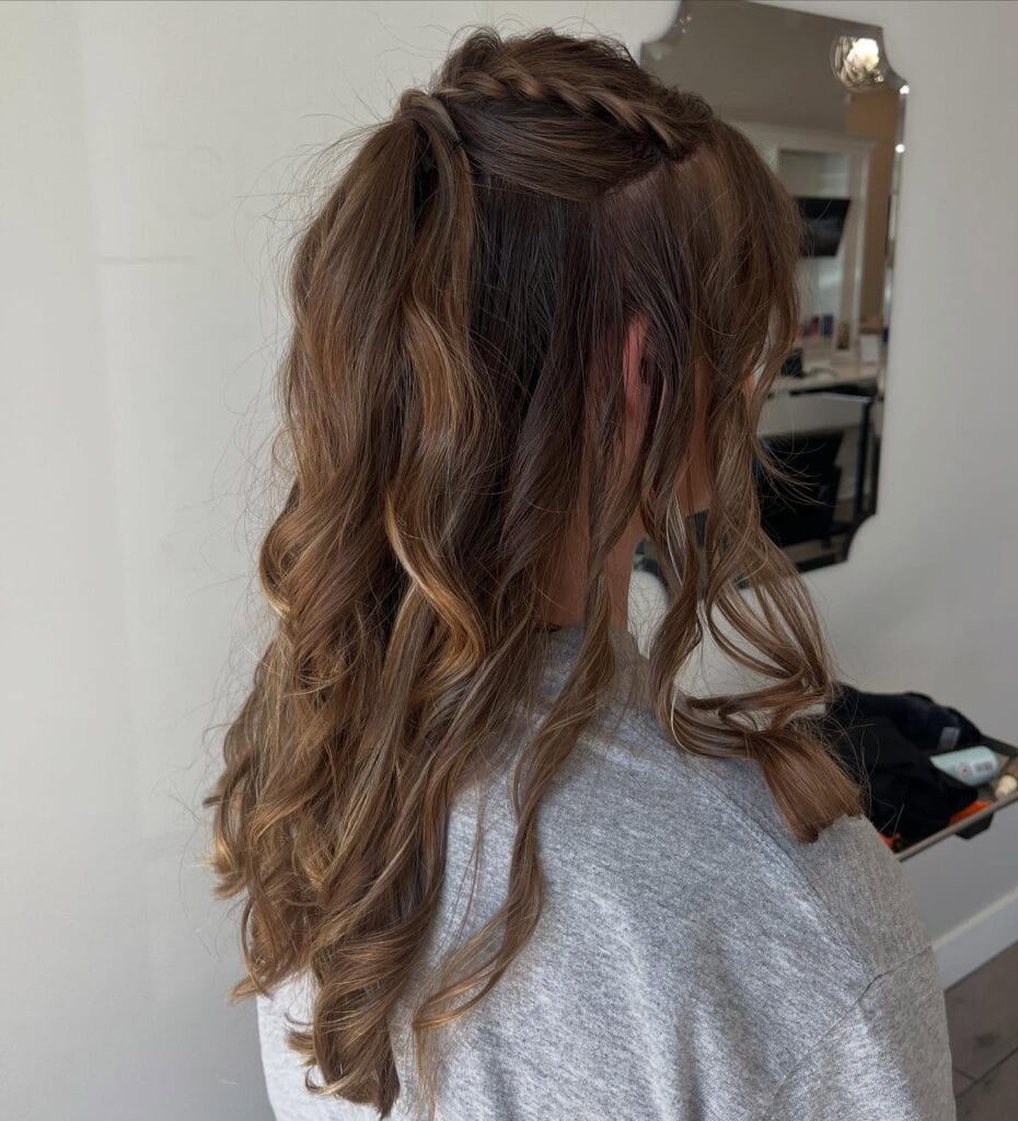 A person with caramel brown hair in a half-up style featuring a crown braid and long loose curls.