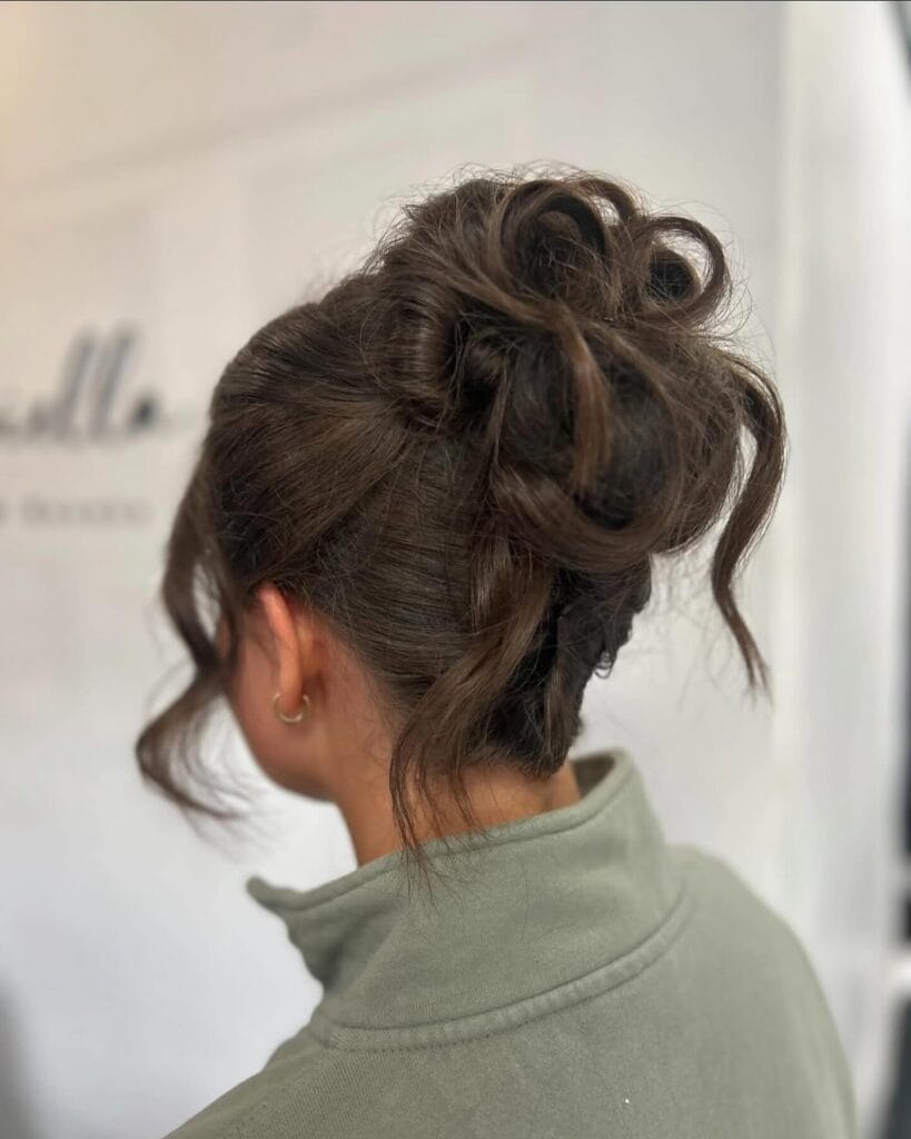 A young woman from behind with dark brunette hair styled in a voluminous, messy high bun with loose wavy tendrils.