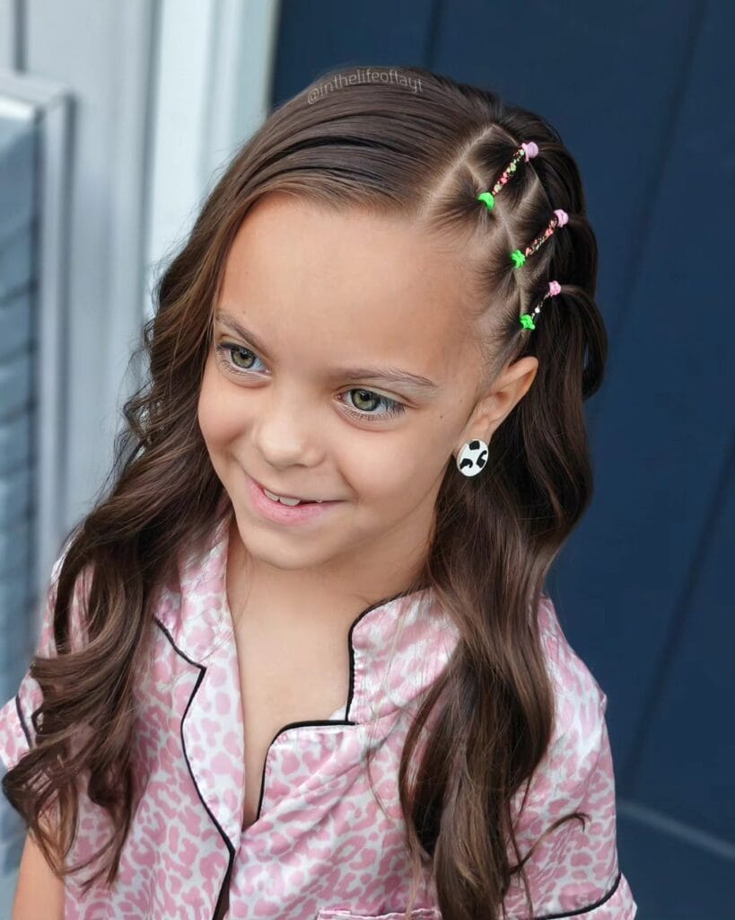 A young girl with long wavy brown hair featuring three small side sections tied with bright pink and green elastics.