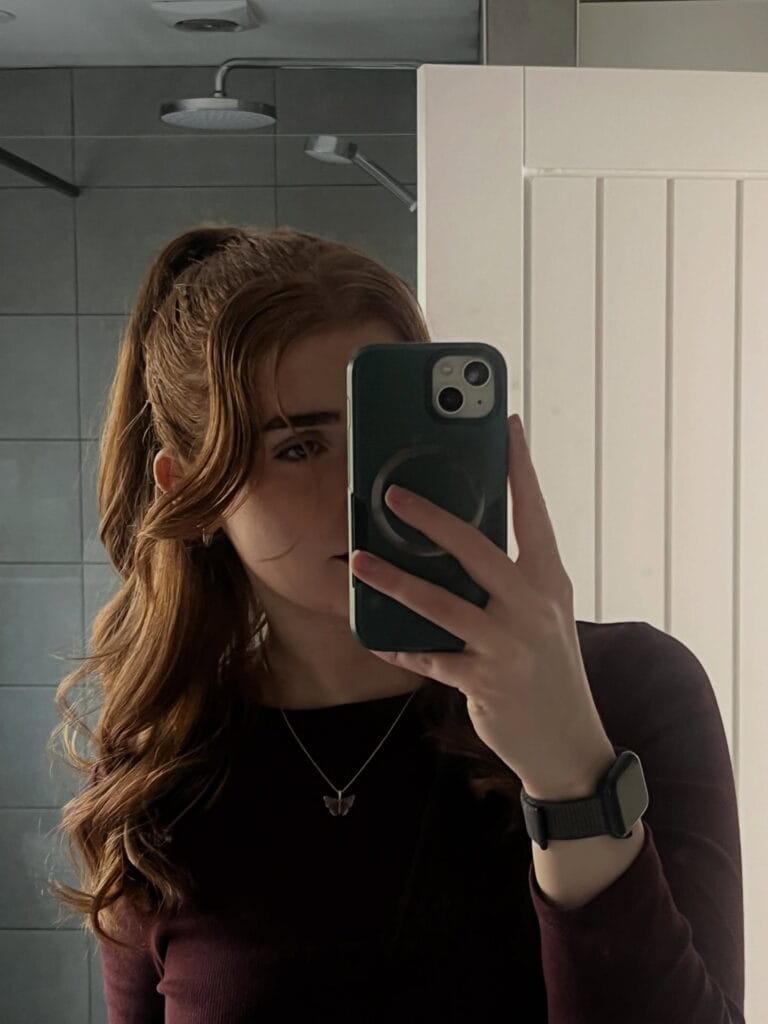 A young woman taking a mirror selfie showing her auburn hair in a high ponytail with wavy, face-framing layers.