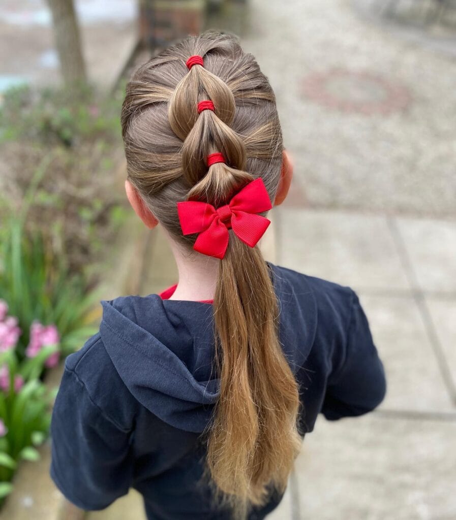 A young girl seen from behind with light brown hair styled in a bubble braid featuring red elastics and a large red bow.
