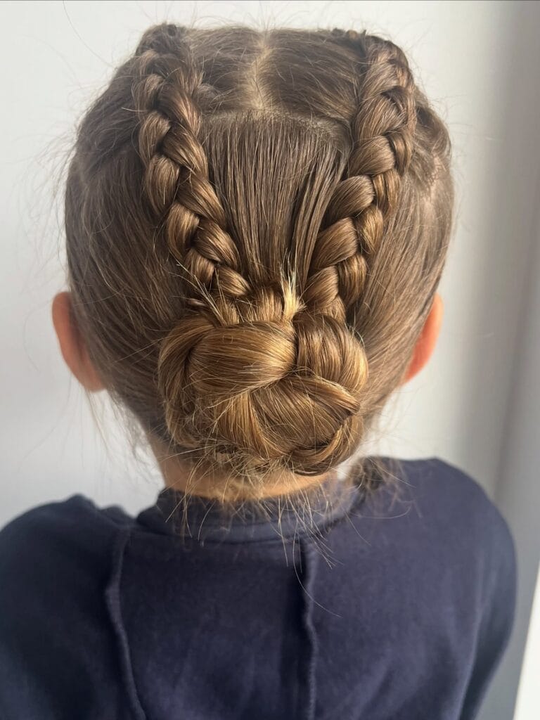 A young girl seen from behind with light brown hair styled into two French braids that lead into a low braided bun.