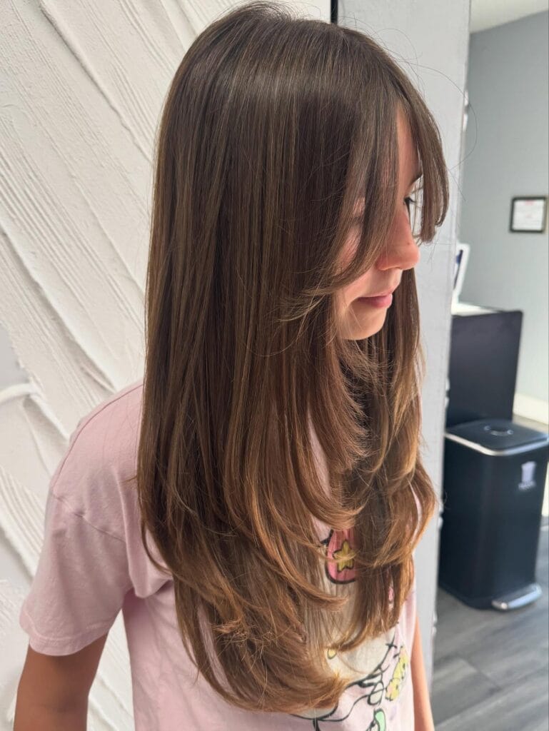 A young woman with long, straight brunette hair featuring soft face-framing layers and curtain bangs.