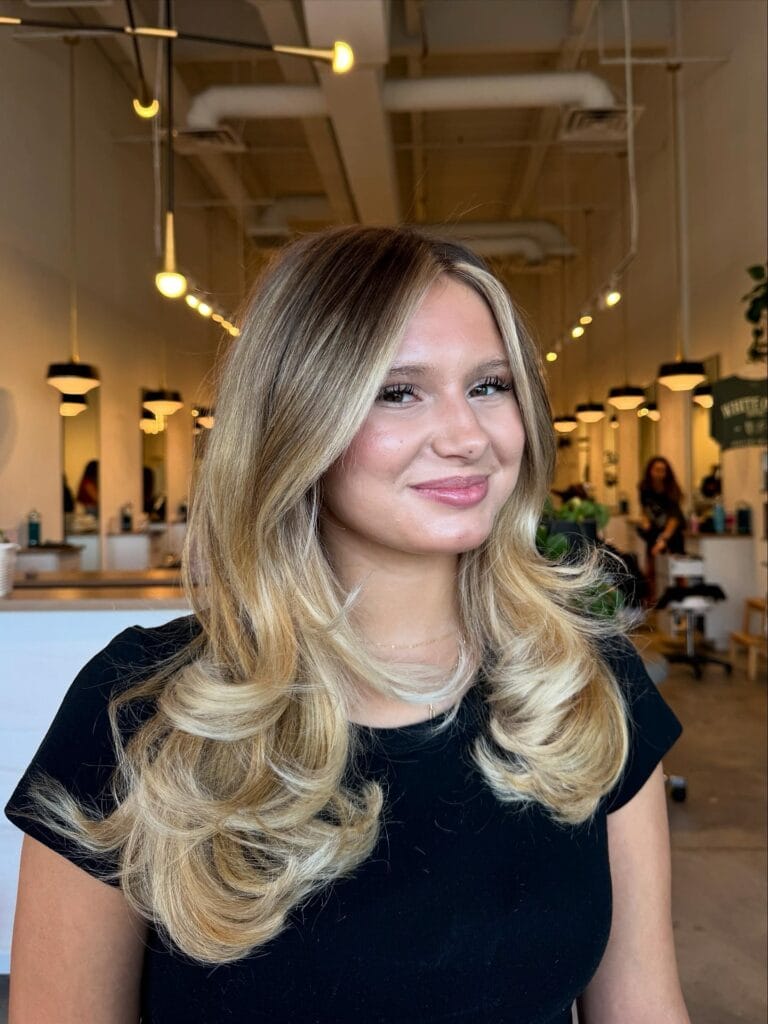 A young woman with layered blonde hair featuring curtain bangs and large, bouncy, flipped-out ends.