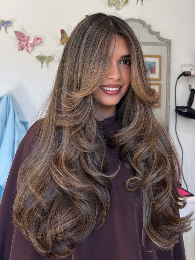 A woman with very long brown hair featuring caramel highlights and voluminous tiered butterfly layers with bouncy curls.