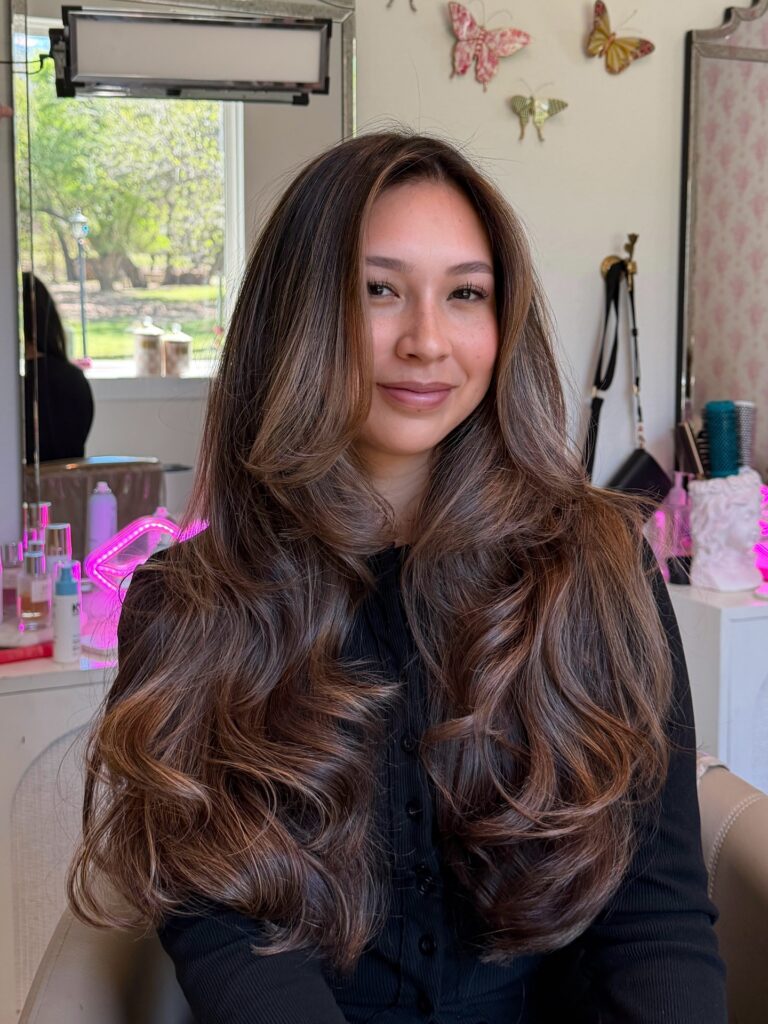A woman with long wavy chestnut brown hair featuring honey highlights and soft layered texture.