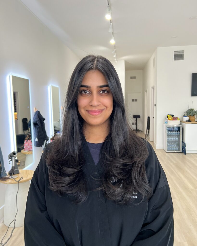 A woman with long glossy jet black hair styled with soft bouncy layers at the ends and delicate face framing pieces.