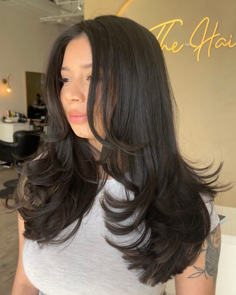 A woman with long thick dark brunette hair featuring voluminous winged layers that flick outward at the ends.