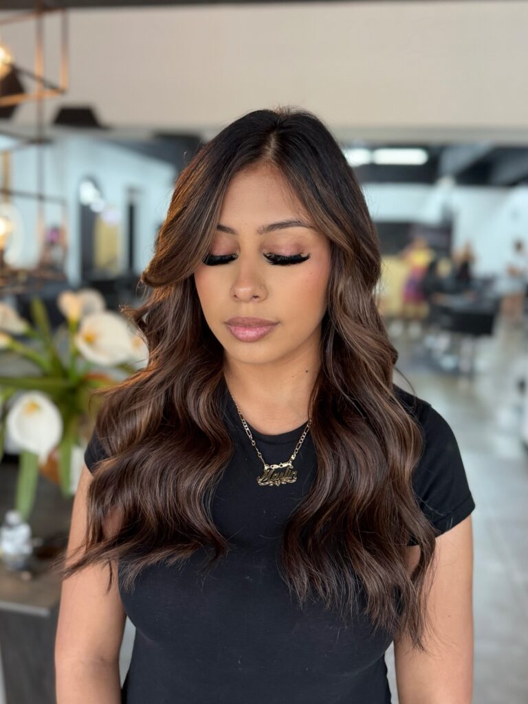 A woman with long dark brown hair featuring caramel balayage and soft beachy waves with face-framing layers.