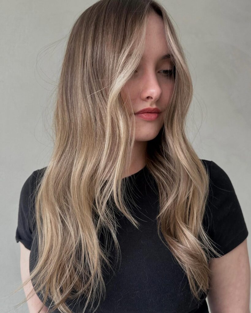A woman with long sandy blonde balayage hair featuring bright face framing highlights and soft layered ends.