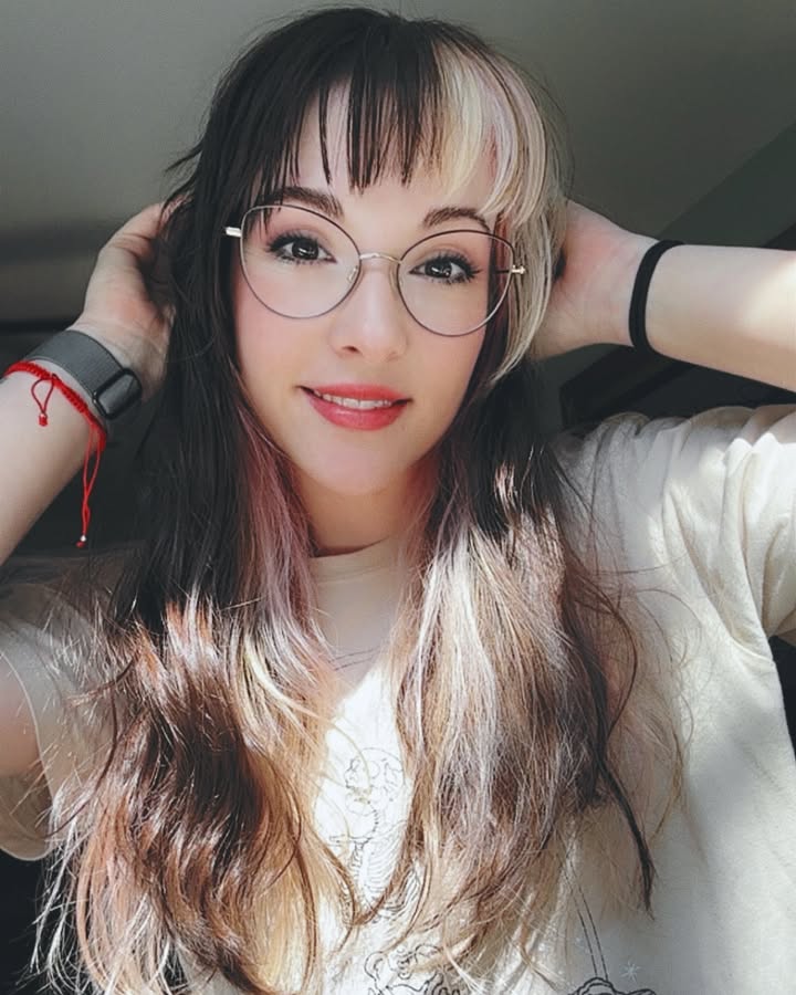 Long two tone layered hair with full soft bangs styled neatly above glasses.