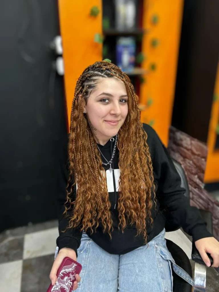 A smiling woman wearing vibrant ginger colored Fulani braids with zigzag partings and long wavy curly ends.