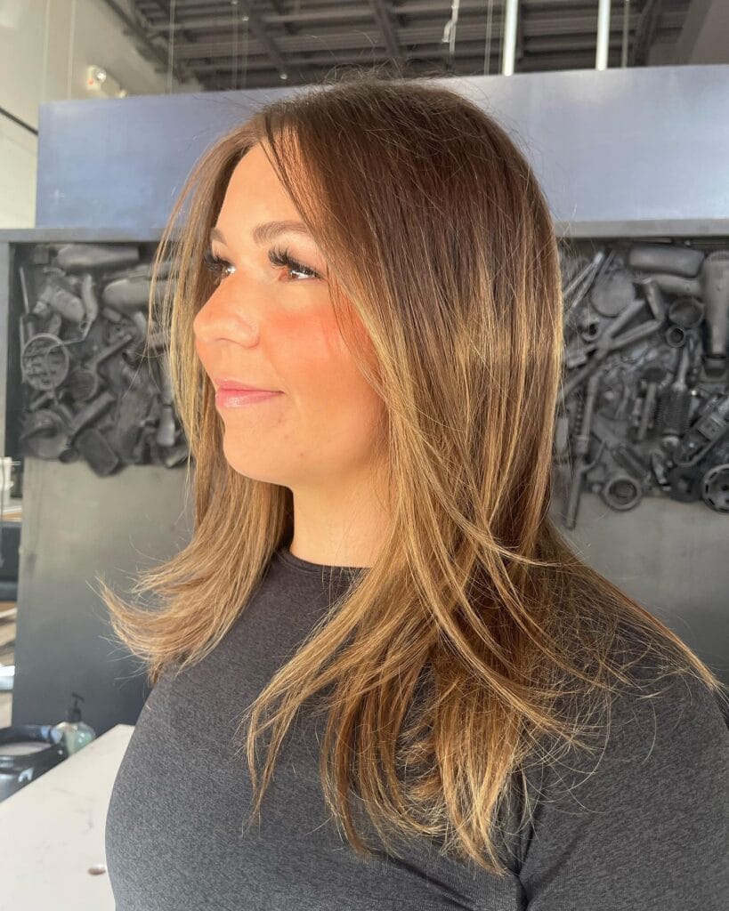 A woman with medium length brown hair featuring honey blonde balayage highlights and face framing layers.