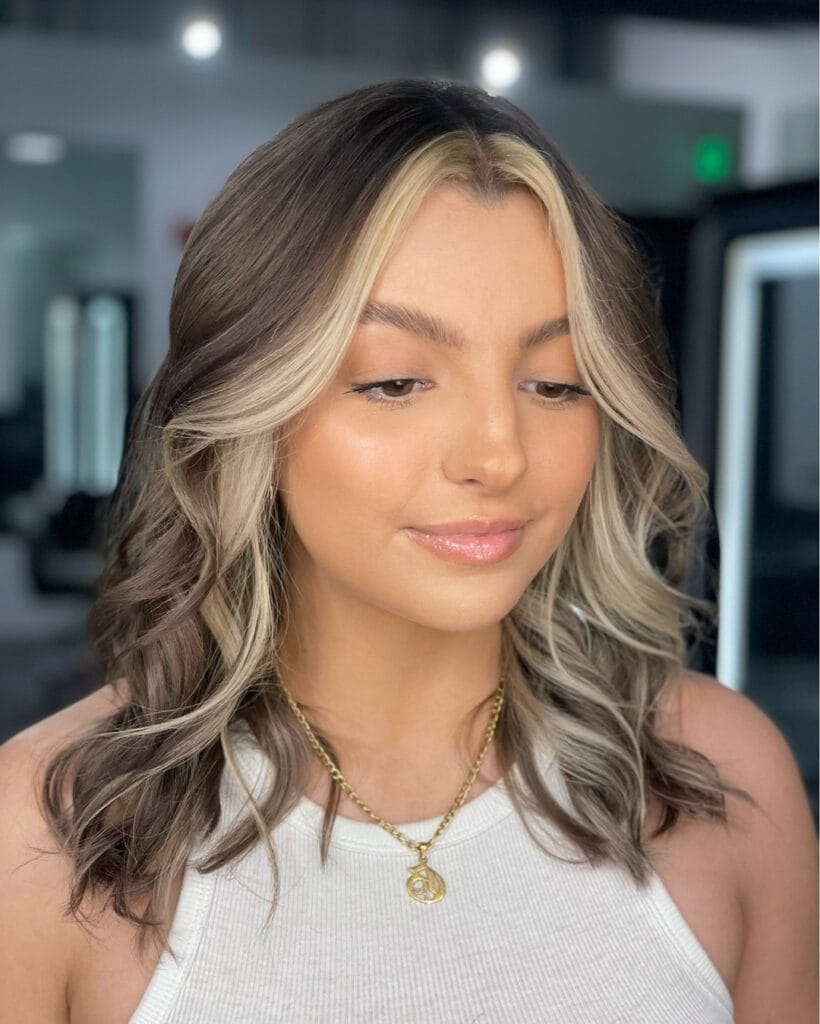 A woman with wavy ashy brunette hair featuring a bold bright blonde money piece highlight around her face.