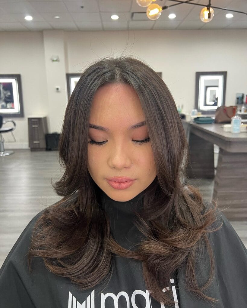 A woman with long dark brown hair styled in voluminous layers and face framing curtain bangs with curled ends.