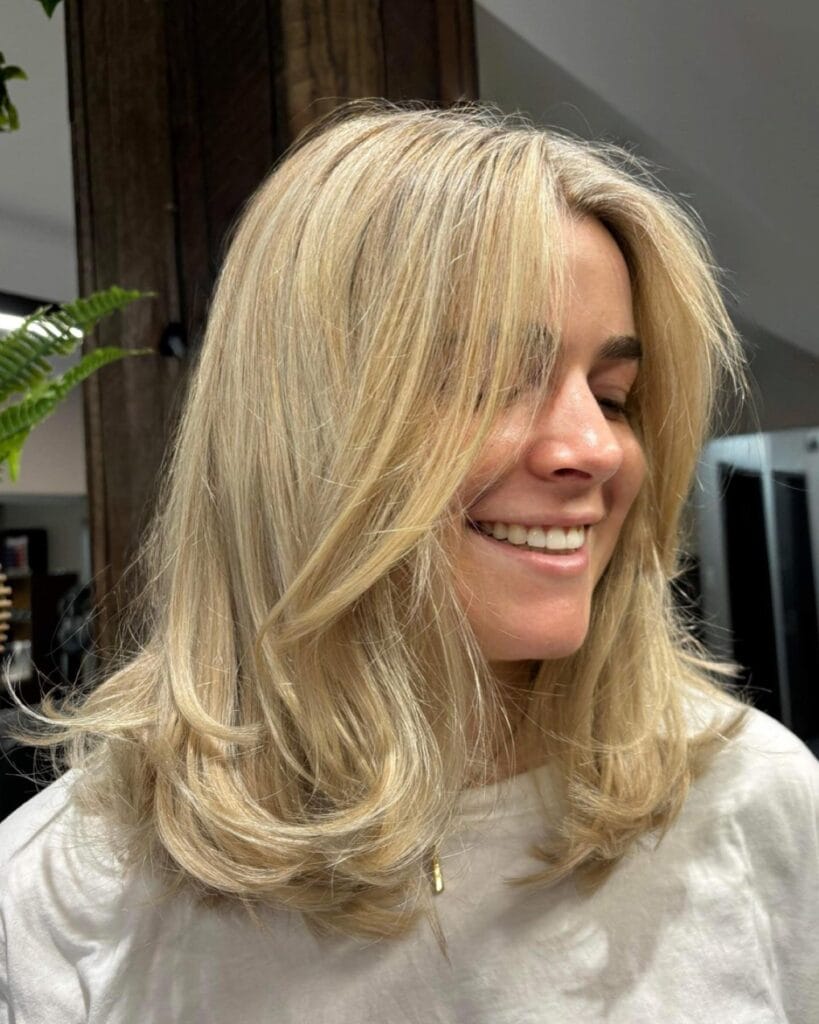 A woman smiling while showing off her medium-length radiant golden blonde hair styled in soft bouncy waves with face-framing layers.