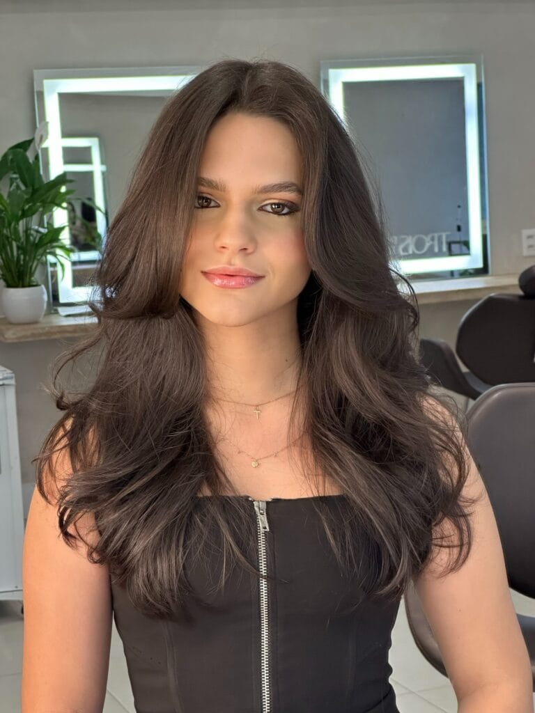 A woman with long dark brown hair featuring layered face framing waves and voluminous undercuts in a beauty salon setting.