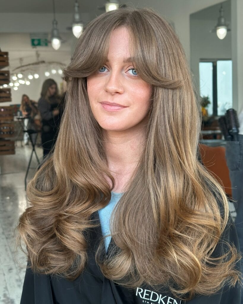 A woman with long honey ash blonde hair featuring voluminous layers and retro flipped bottleneck bangs.