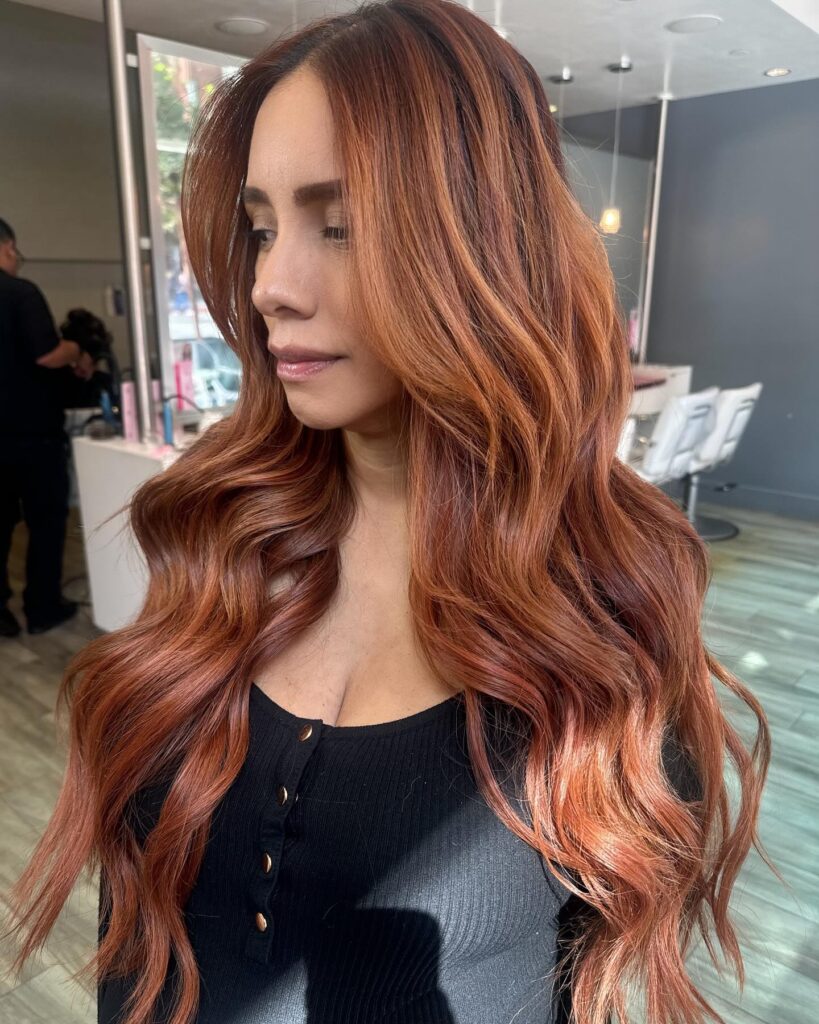 A woman with long vibrant copper red hair styled in large glossy waves and voluminous layers.
