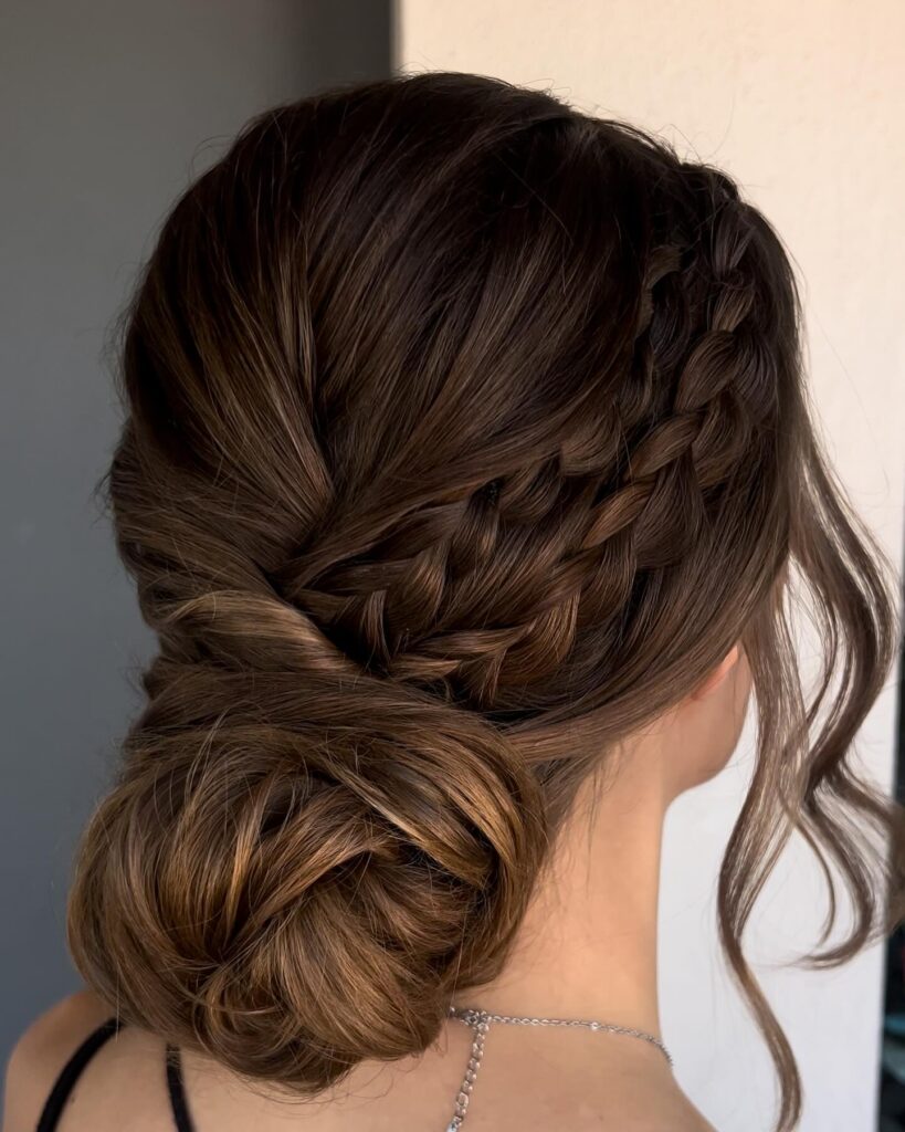 A woman with deep brown hair styled in a side braid that leads into a large textured low bun.