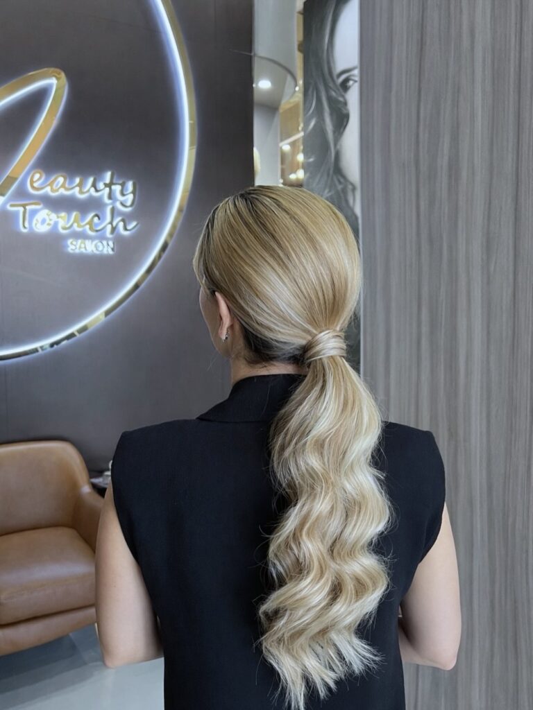 A woman with long wavy blonde hair styled in a sleek low ponytail with a hair wrap at the base.