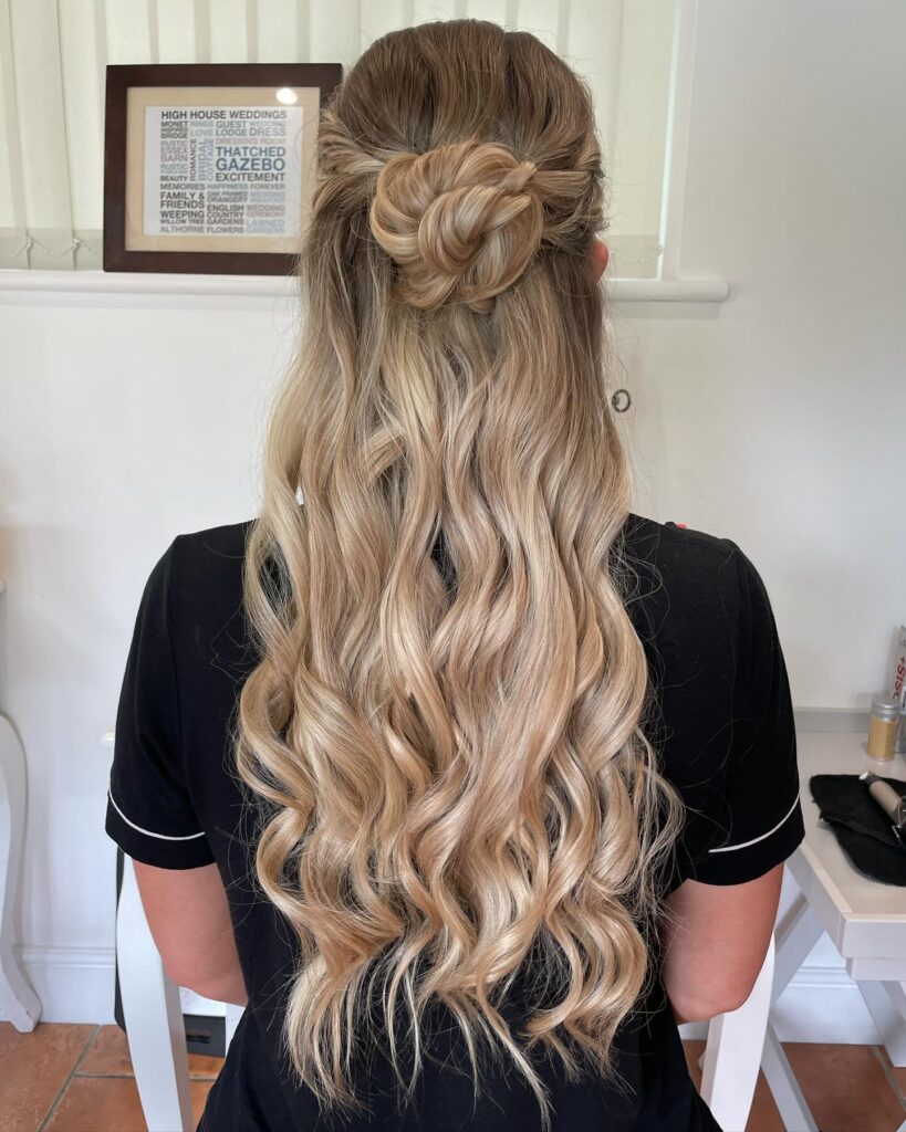 A woman viewed from the back has long blonde hair styled with a rose shaped half up knot and flowing defined waves.