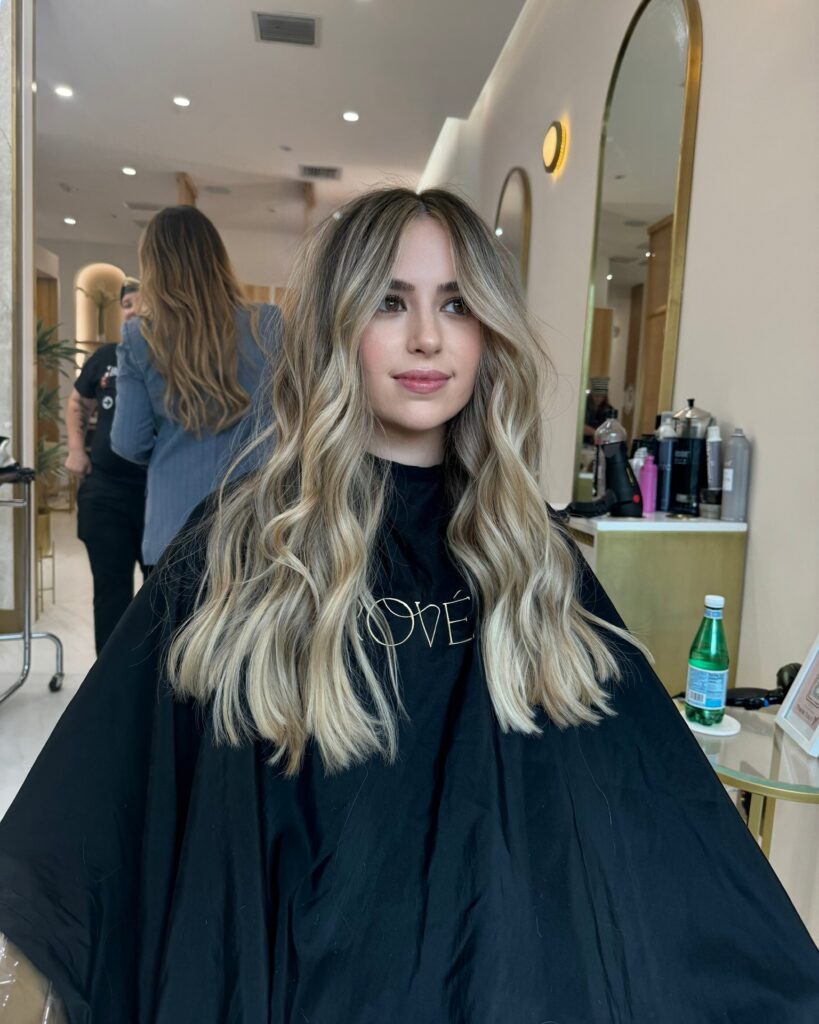 A woman with long sun kissed balayage hair styled in voluminous beachy waves with a center part.