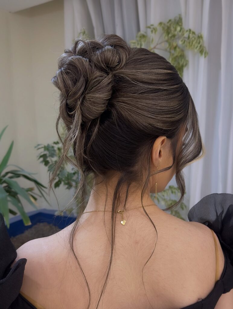A woman showcases a sophisticated low bun with intricate textured loops and ash blonde highlights with soft tendrils framing her face.