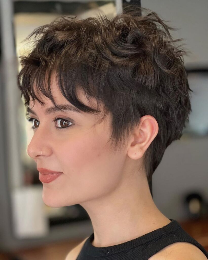A short layered tomboy pixie haircut with textured volume on top and soft wispy fringe in a natural brunette shade.