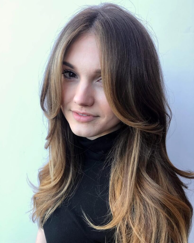 A woman with long layered brown hair with soft waves and subtle face framing highlights.