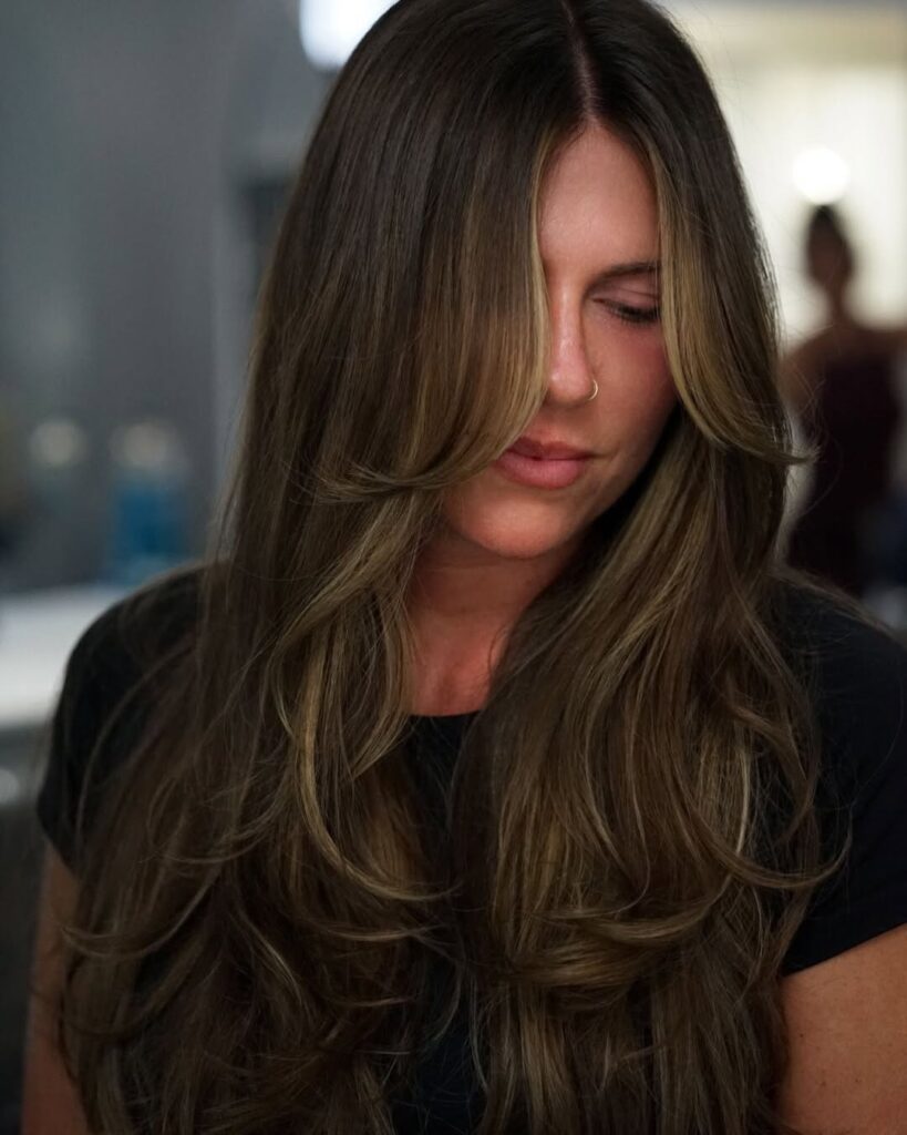 A woman with long dark brown layered hair styled in voluminous waves.