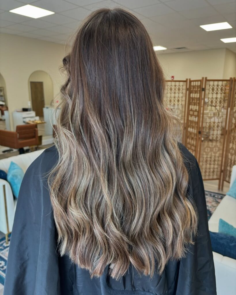 A woman with long layered blonde balayage hair styled in loose waves.