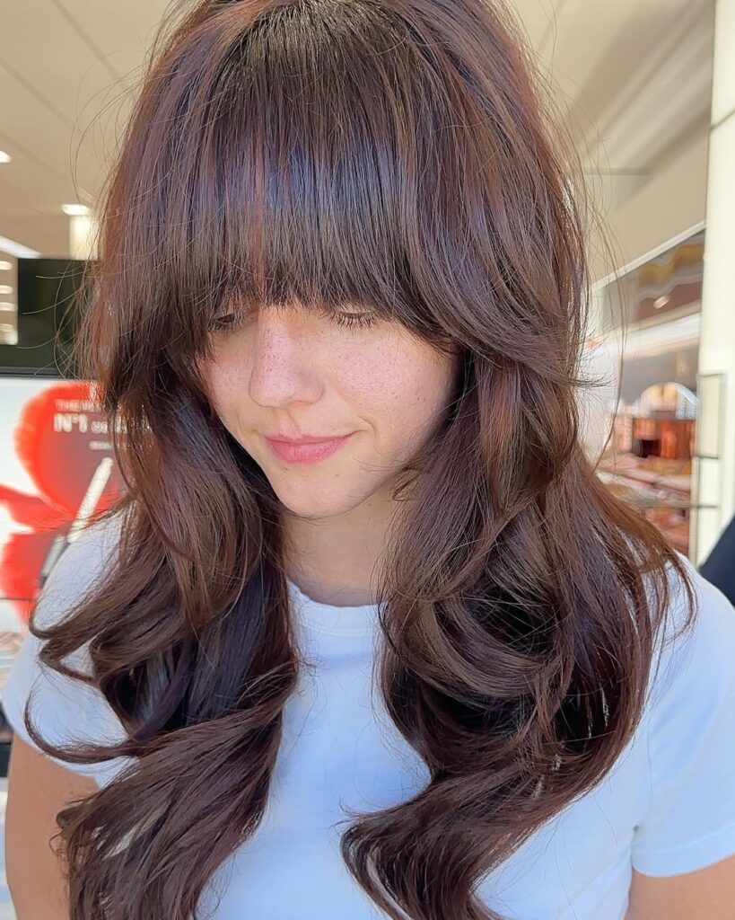 A woman with long layered brunette hair styled in soft waves with a full fringe.