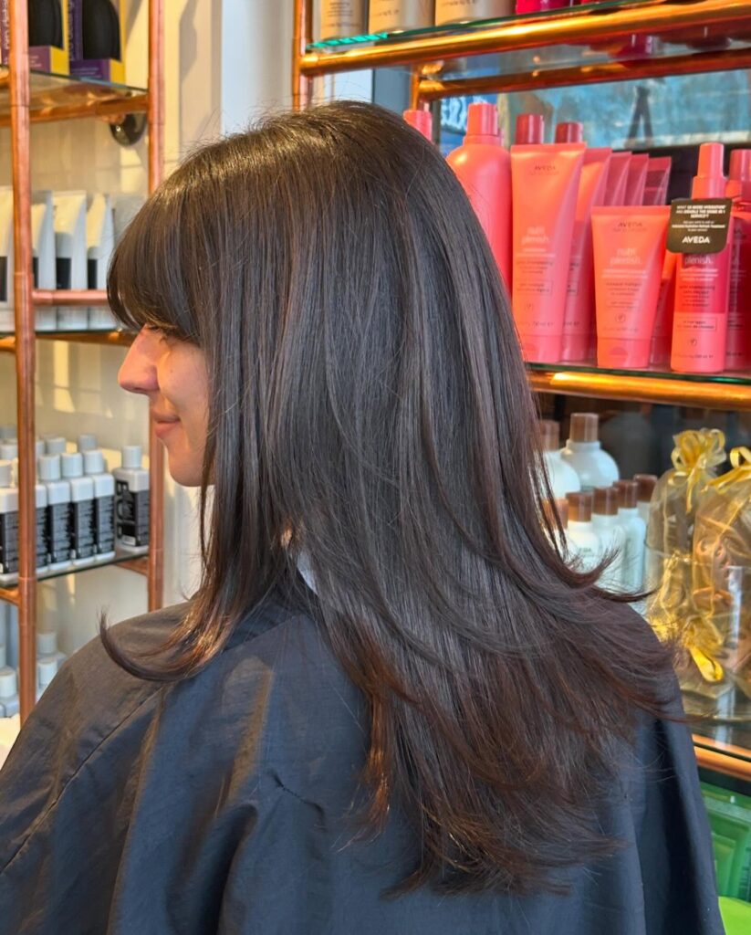 A woman with long glossy espresso brown hair featuring soft wispy bangs and subtle tiered layers at the ends.