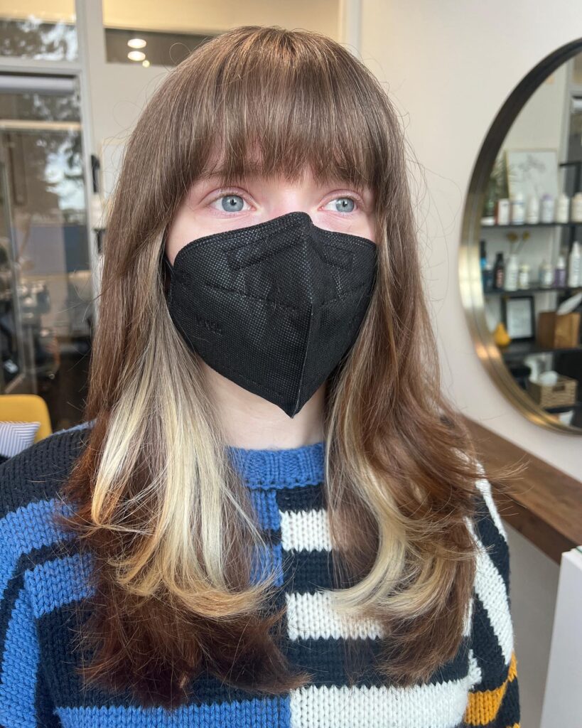 A woman with medium length brown hair and full bangs featuring bright blonde peekaboo highlights underneath the layers.