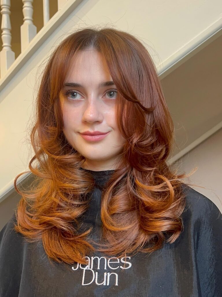 A woman with medium length auburn hair styled in voluminous bouncy layers and soft curtain bangs.