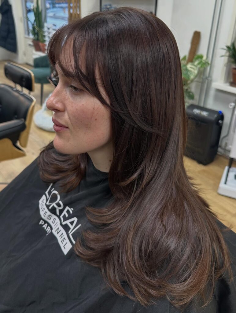 A woman with long dark espresso brown hair featuring soft wispy bangs and tiered layers with curved ends.