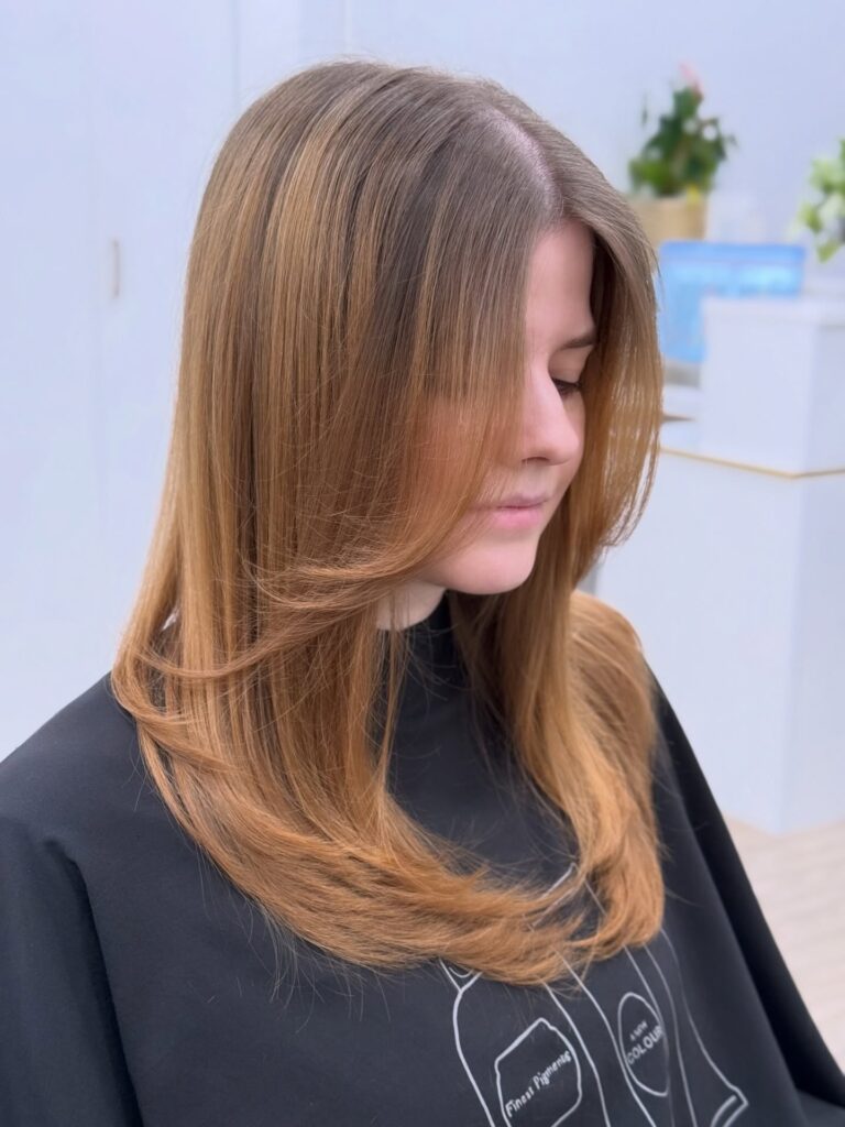A woman with long honey blonde hair featuring face framing butterfly layers and soft outward curls at the ends.